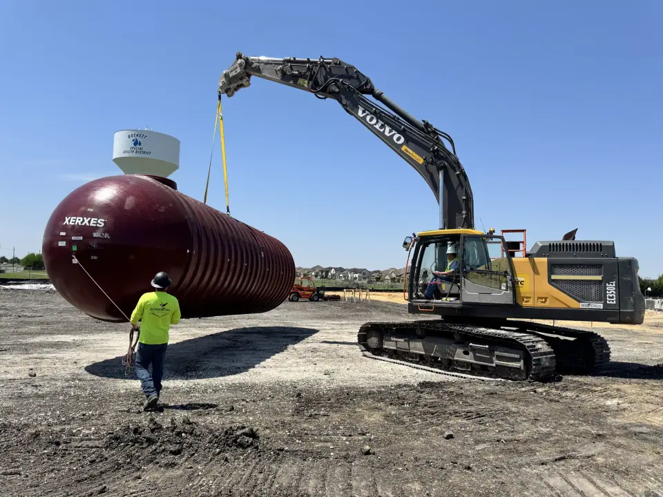 Crew guiding a tank with a tagline as the excavator moves it